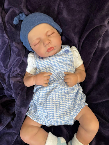 Sleeping reborn baby doll wearing blue knit hat, blue checkered romper, and white socks on purple blanket – realistic handmade newborn doll